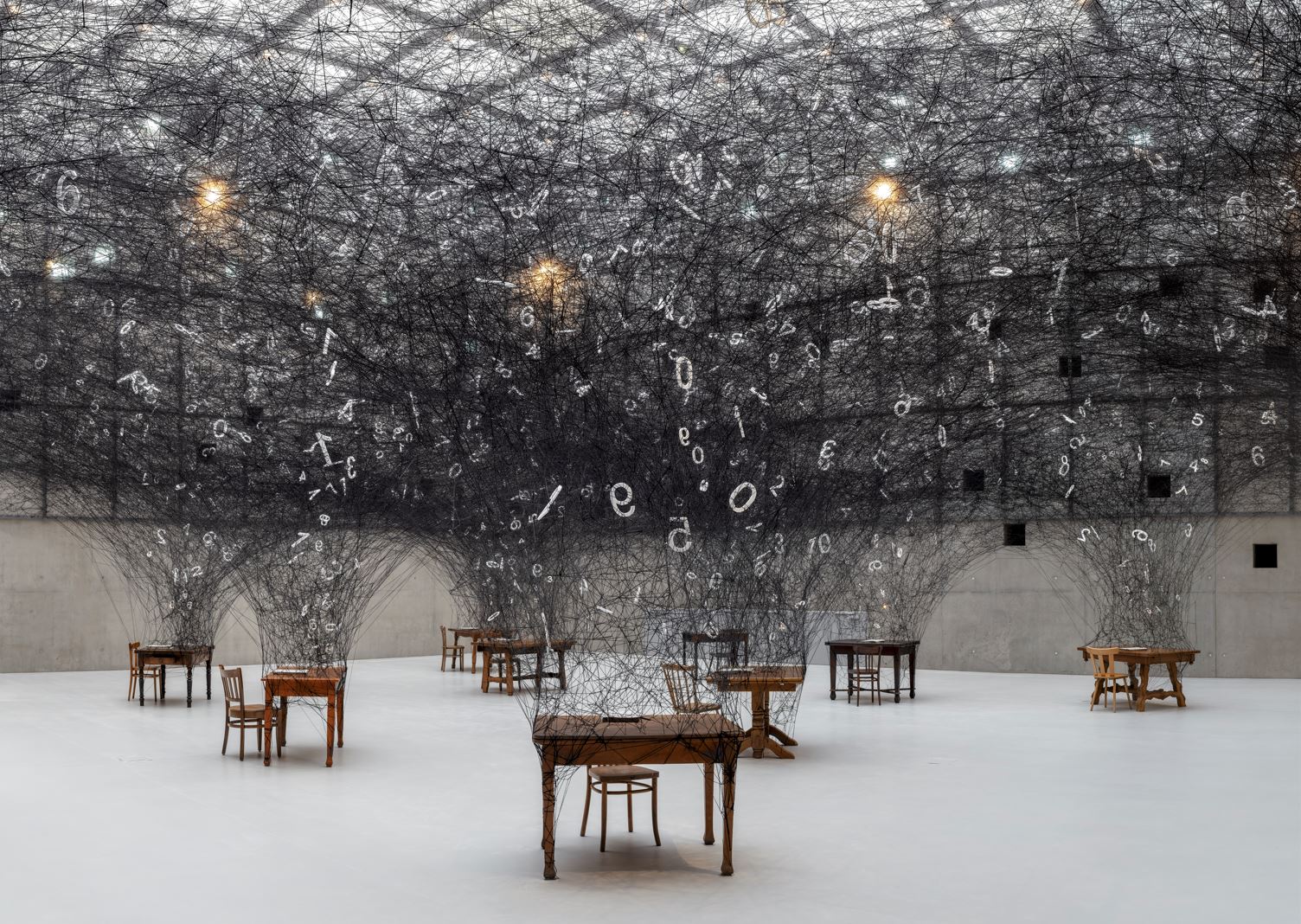 Art installation with black threads and suspended numbers and letters over wooden tables.